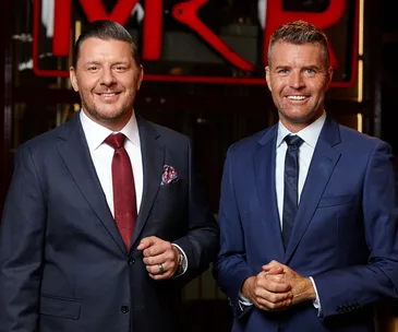 Two men in suits smiling in front of a "MKR" sign, possibly judges or hosts related to a TV show.