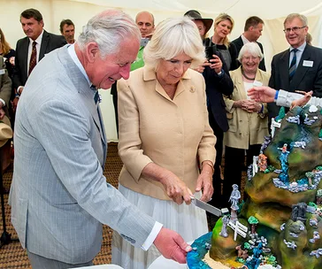 Royal couple cutting an elaborate multi-tiered birthday cake with decorative landscape elements, surrounded by onlookers.