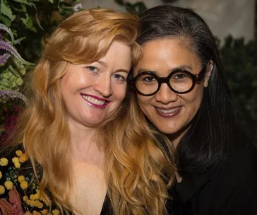Two women smiling closely together at an event, one with long red hair, the other with black hair and round glasses.