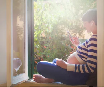 Pregnant woman in striped shirt listening to a podcast with earphones, sitting by a sunny window holding a smartphone.