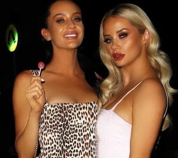 Two women posing together at night; one holds a lollipop and wears a leopard-print dress, the other wears a light-colored top.