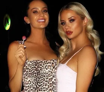 Two women posing together at night; one holds a lollipop and wears a leopard-print dress, the other wears a light-colored top.