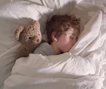 A child sleeps peacefully in bed, snuggled under white blankets with a teddy bear beside them.
