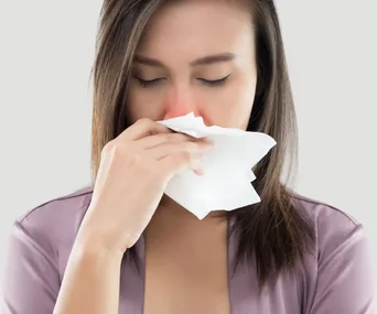 Woman with closed eyes holds tissue to nose, indicating cold or allergy.