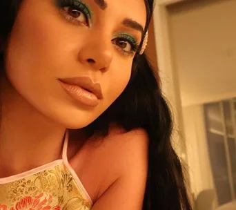 Portrait of a woman with long dark hair, wearing a floral top and blue eyeshadow, posing indoors.