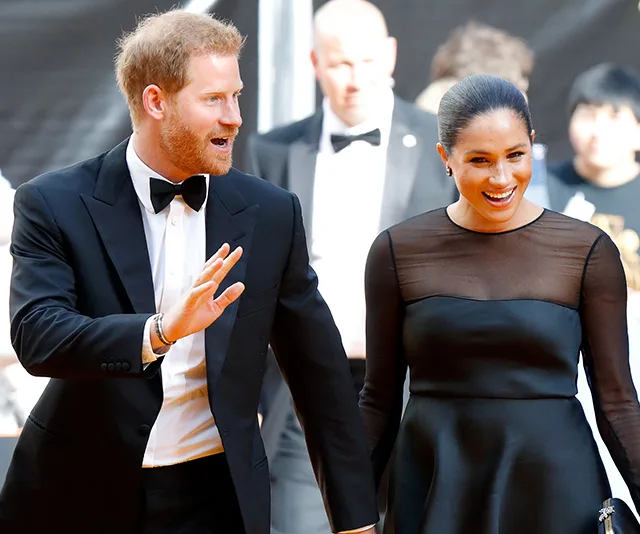 Prince Harry and Meghan Markle smile and wave on the red carpet, dressed in formal attire.