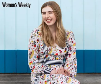 A woman in a floral dress smiles against a light blue wooden backdrop, logo "Women's Weekly" visible.