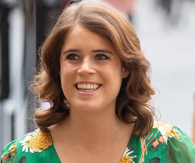 A woman with wavy hair wearing a vibrant green dress with floral patterns smiles while standing outside.