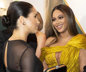 Two women smiling at each other at an event; one in black and the other in a yellow dress.