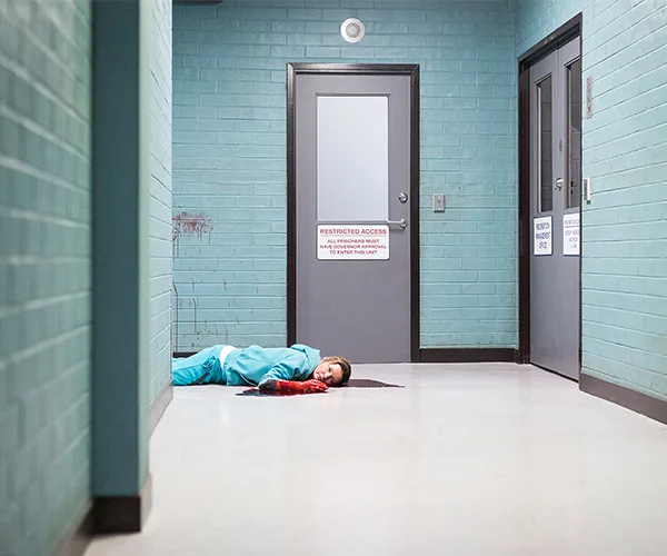 Prison corridor scene with a person in a blue uniform lying on the ground, a wound and blood visible, near restricted access doors.