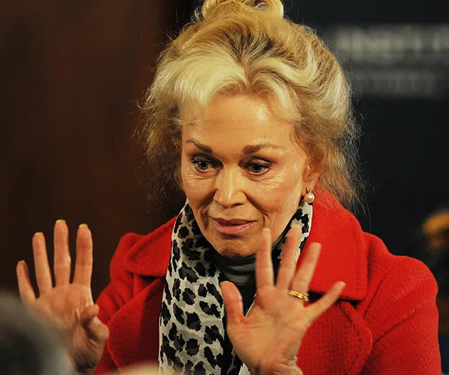 An older woman with blonde hair gestures with her hands in a leopard print scarf and red coat.