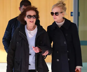 Two women walking together, both wearing sunglasses and coats, with a man in the background.