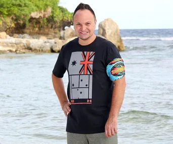 Man standing on a beach wearing an Australian-themed shirt and a "Survivor" armband, with ocean and rocks in the background.