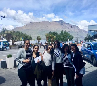 Group of six people smiling outdoors with a mountain backdrop and blue cars parked nearby under a partly cloudy sky.