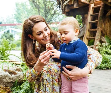 A woman in a floral dress shows a small object to a toddler in a blue sweater in a garden setting.