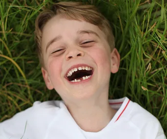A young boy with light hair laughs joyfully lying on grass, wearing a white shirt.