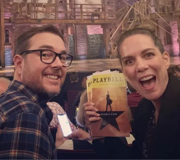 A couple smiling with a "Hamilton" Playbill in hand, seated in a theater with the stage visible in the background.