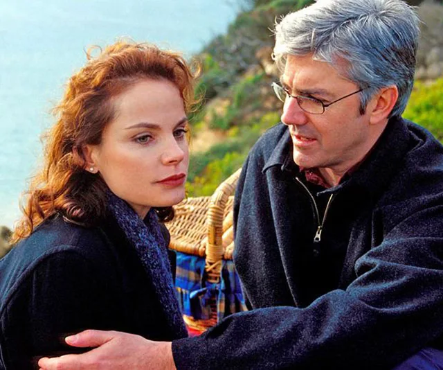 A man and woman sit by the sea with a picnic basket, engaged in a serious conversation.