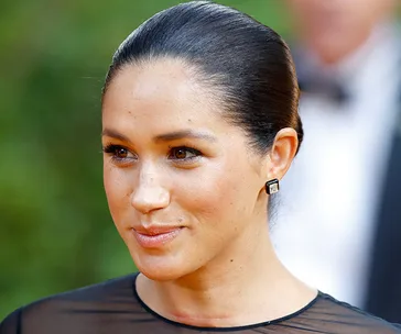 Meghan Markle with slicked back hair, wearing elegant earrings and a sheer top, smiling outdoors.
