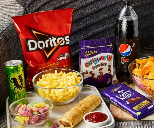 Snack assortment: Doritos, curly fries, Cadbury chocolates, Pepsi Max, energy drink, and a spring roll with sauce.