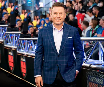 Man in checkered blue suit standing by a railing, smiling, with an audience in the background at an event.