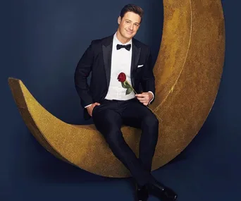 Man in a tuxedo sits on a golden crescent moon, holding a red rose, against a blue background.