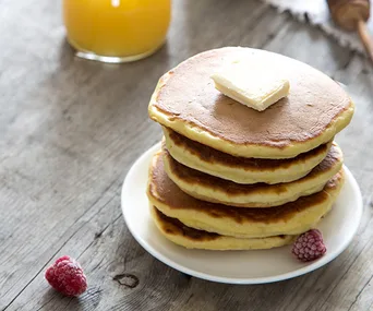 Stack of keto pancakes with butter on top, served on a plate with raspberries; orange juice in the background.