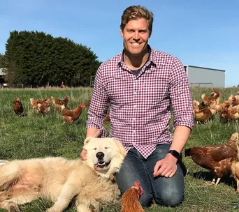 Man smiling with a white dog in a field, surrounded by chickens.