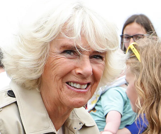 Camilla in a trench coat, smiling outdoors with people in the background.