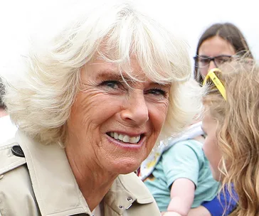 Camilla in a trench coat, smiling outdoors with people in the background.