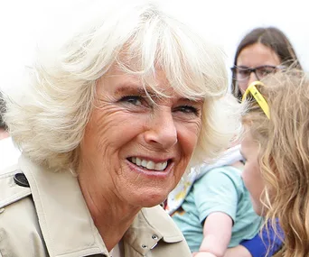 Camilla in a trench coat, smiling outdoors with people in the background.