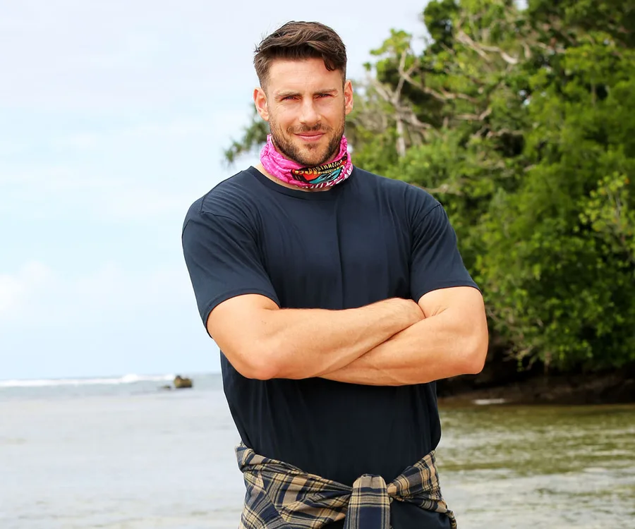 Man standing by the beach with arms crossed, wearing a colorful neck buff, in a lush green setting.