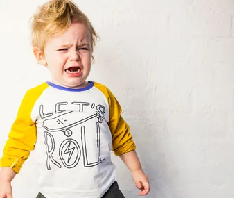 Young child crying, wearing a "Let's Roll" shirt with yellow sleeves, standing against a plain white wall.