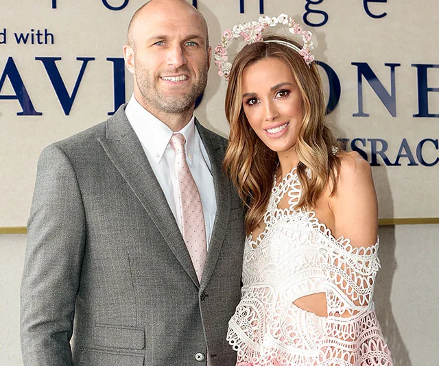 A smiling couple dressed elegantly stands in front of a branded backdrop featuring floral headpiece detail.