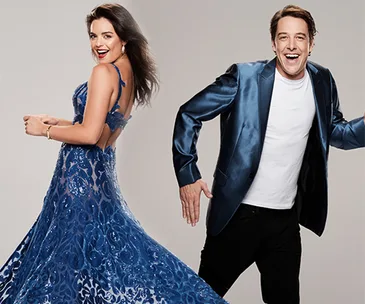 Two people smiling, the woman in a blue gown and the man in a blue jacket and white shirt, dancing enthusiastically.