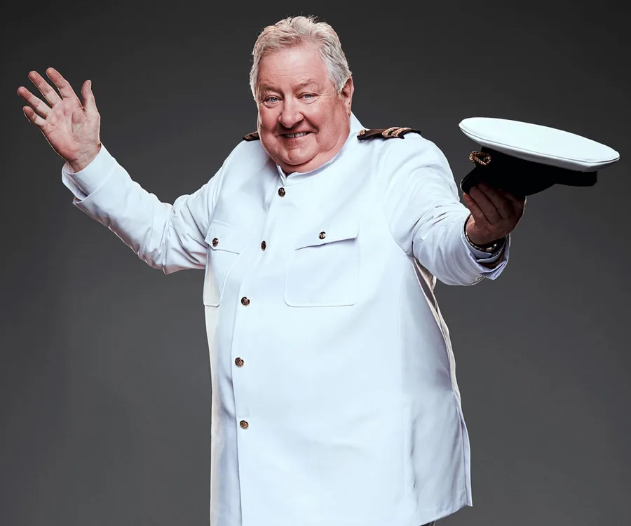 Man in a white naval uniform smiling, holding a captain's hat, with arms open wide against a dark background.