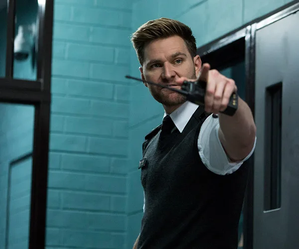 Security officer in a uniform holds a walkie-talkie in a tense moment inside a teal-painted building.