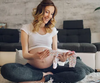 Pregnant woman sitting on a carpet, smiling at a small piece of baby clothing in her hands.