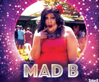Drag queen Mad B posing playfully in a sparkly pink background, wearing a red dress and floral hair accessory.