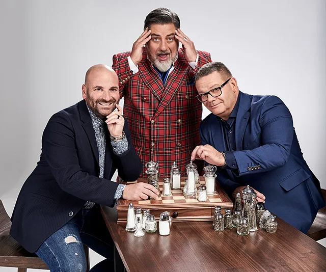 Three men, dressed smartly, playfully pose with a chessboard made of salt and pepper shakers.