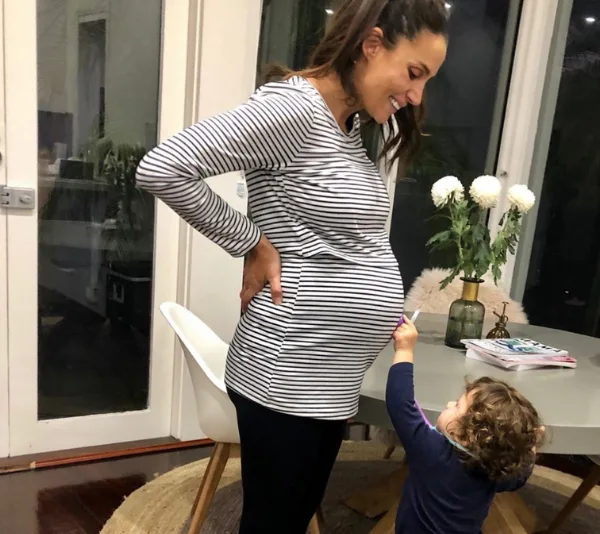 Pregnant woman in striped shirt smiling down at a small child pointing at her belly in a cozy home setting.