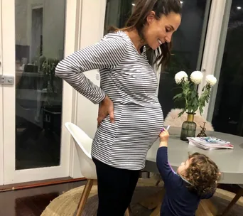 Pregnant woman in striped shirt smiling down at a small child pointing at her belly in a cozy home setting.