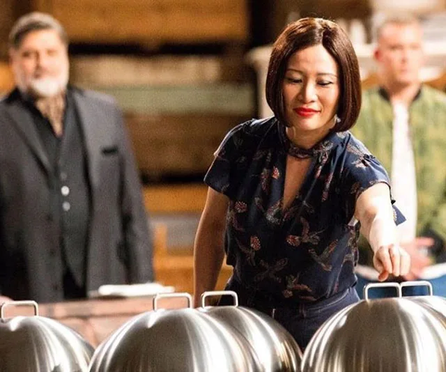 Judge Poh Ling Yeow lifts a cloche on MasterChef set, with two men in the background watching.