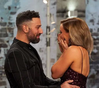 A couple embraces with joy, the man smiling, and the woman covering her mouth in surprise, against a blurred background.