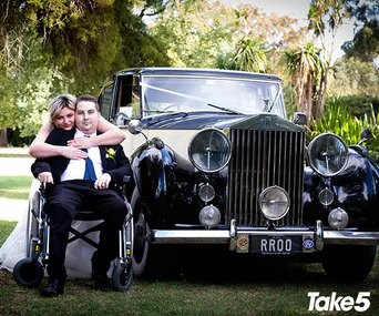 Bride embraces groom in wheelchair in front of vintage car; greenery in background.