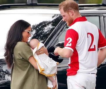A woman holds a baby while a man in a red and white polo shirt stands nearby, smiling, next to a car.