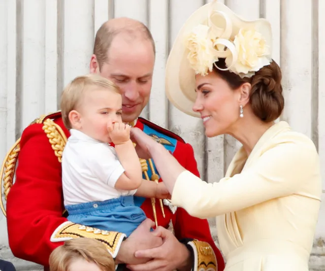 Family gathering with a man in red uniform holding a child, and a woman in a yellow dress with a floral hat smiling.