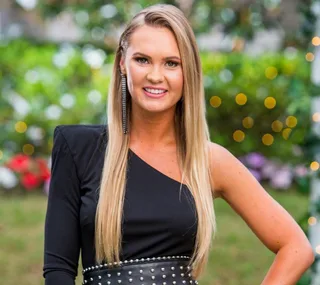 Woman in a black one-shoulder dress with long blond hair, smiling outdoors with greenery and lights in the background.