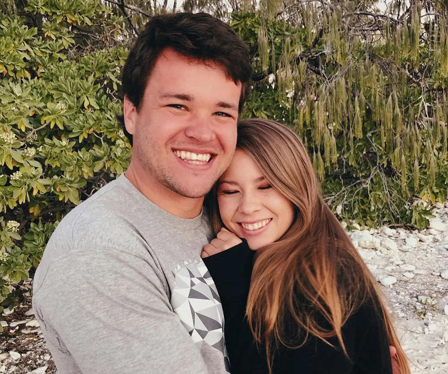 A couple smiling and embracing outdoors with greenery in the background.
