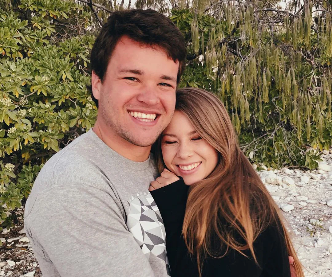 A couple smiling and embracing outdoors with greenery in the background.